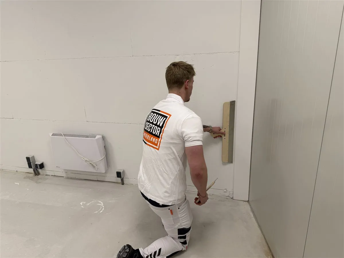 Stukadoor Bram van Bouwsector Nederland zet knielend een spaan tegen de onderkant van de wand