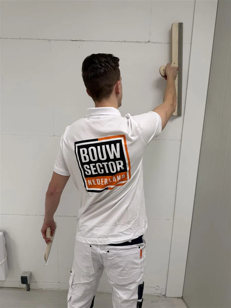 Vakman Argi van Bouwsector Nederland houdt een spaan tegen de wand, met het logo op zijn rug